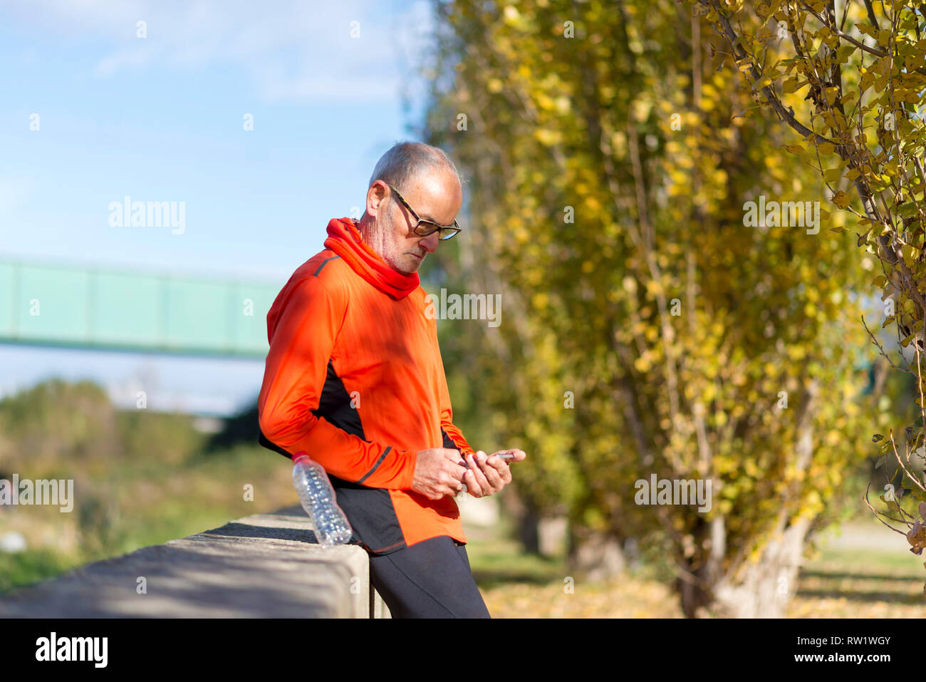 Vue latérale d'un coureur senior man leaning on fence lors du test de l'exercice dans un téléphone mobile à l'extérieur par une journée ensoleillée Banque D'Images