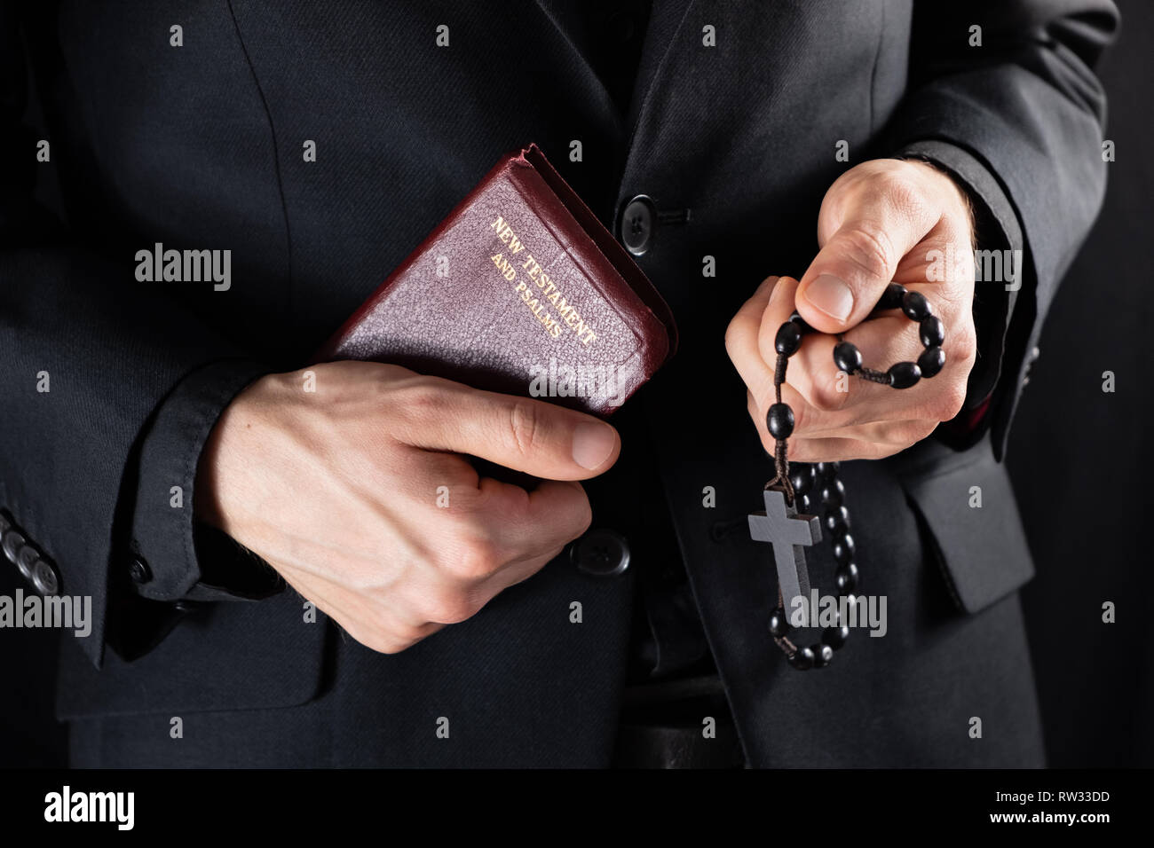 Mains d'un prêtre chrétien vêtus de noir tenant un crucifix et livre du Nouveau Testament. Personne religieuse avec Bible et chapelet, low-key image. Banque D'Images
