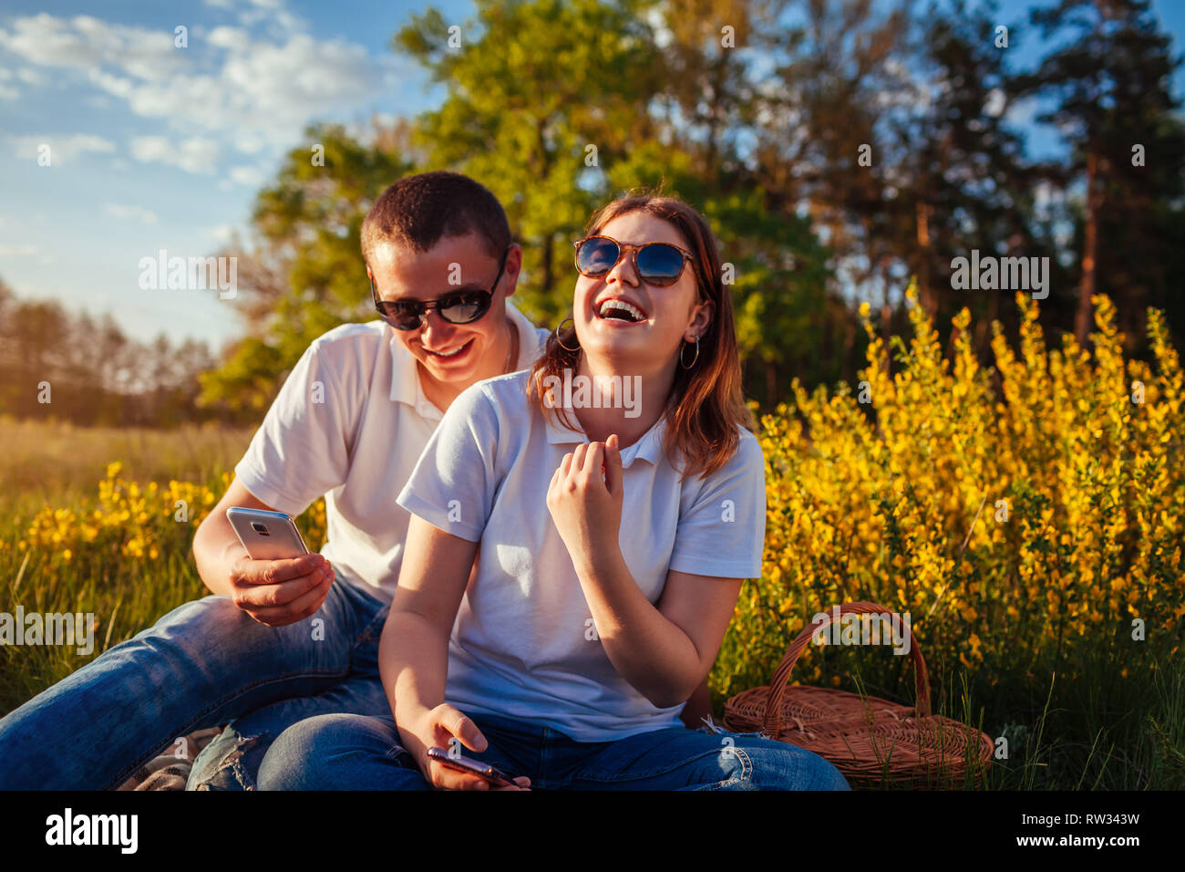 Happy young couple accroché dans leurs téléphones et de rire à l'extérieur. La femme et l'homme se détendre après avoir pique-nique au coucher du soleil. Banque D'Images