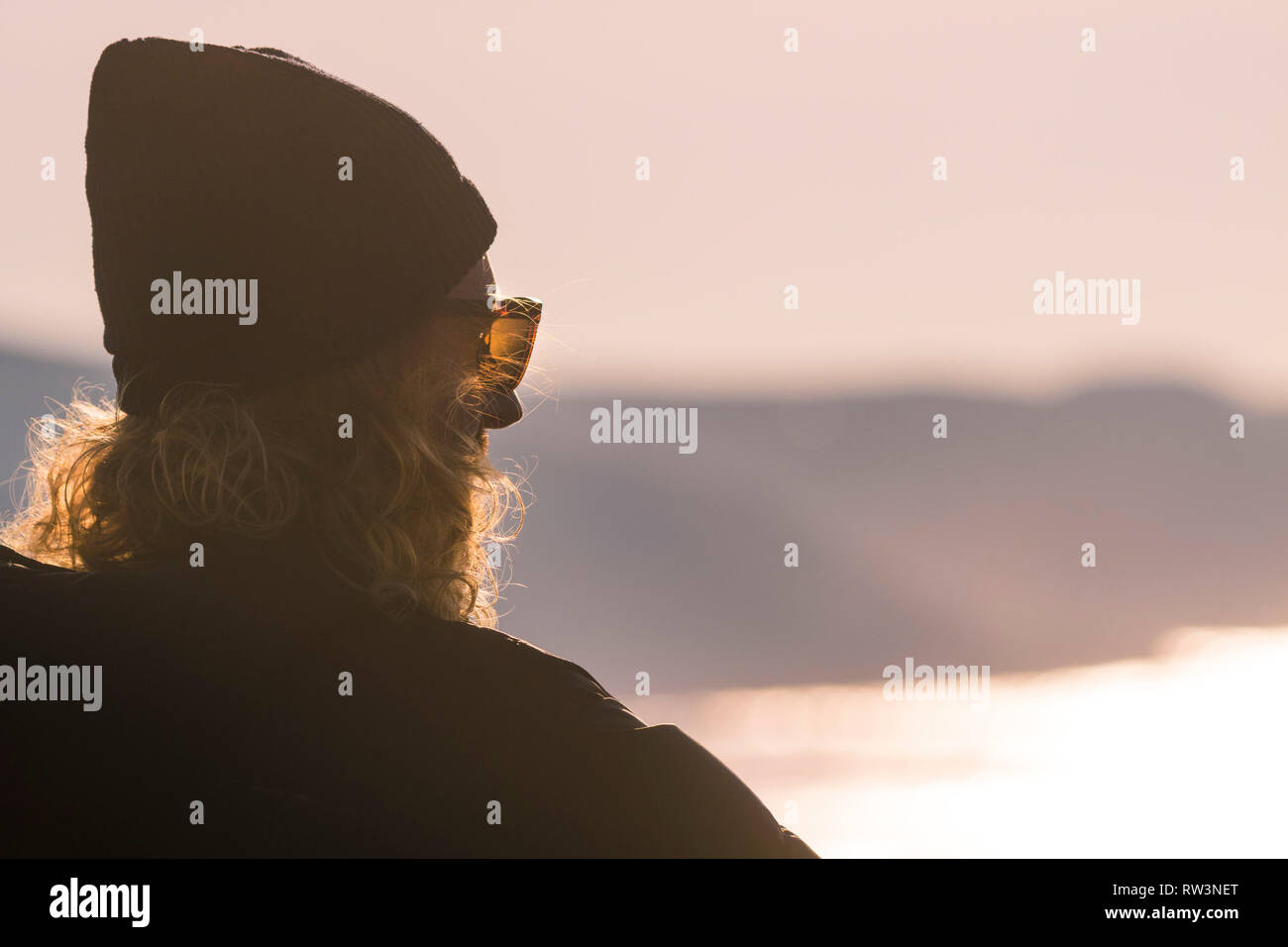 Un homme avec de longs cheveux blonds et portant un bonnet et lunettes de soleil face à la mer. Banque D'Images