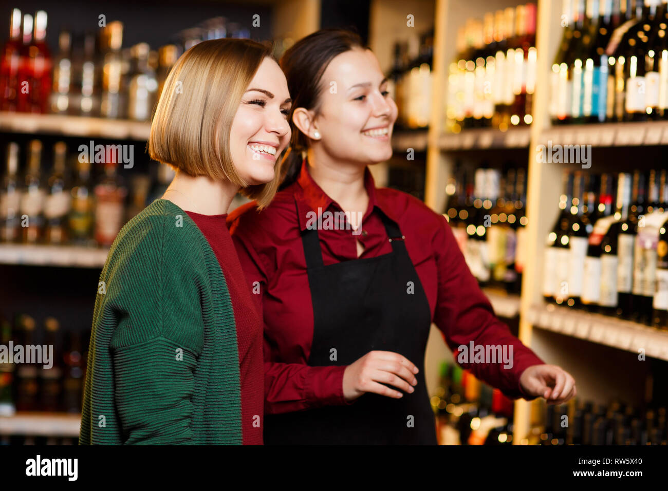 Photo de deux femmes souriant sur fond flou d'étagères avec des bouteilles de vin en magasin Banque D'Images