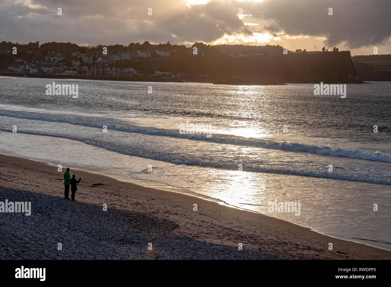 Ballycastle, Irlande du Nord, Royaume-Uni. Côte de l'Atlantique. Plage, vagues et méconnaissable paire dans la réflexion de la lumière au coucher du soleil avec des nuages lourds Banque D'Images