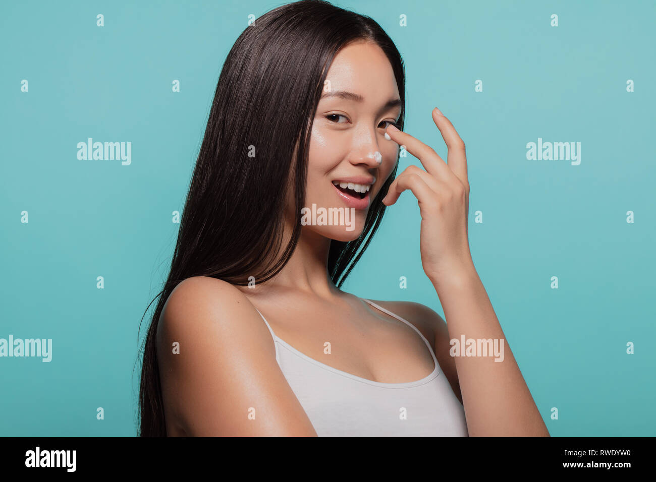 Modèle féminin asiatique l'application de l'hydratant sur son nez et en regardant la caméra. Woman applying moisturizer cream sur son visage contre fond bleu. Banque D'Images