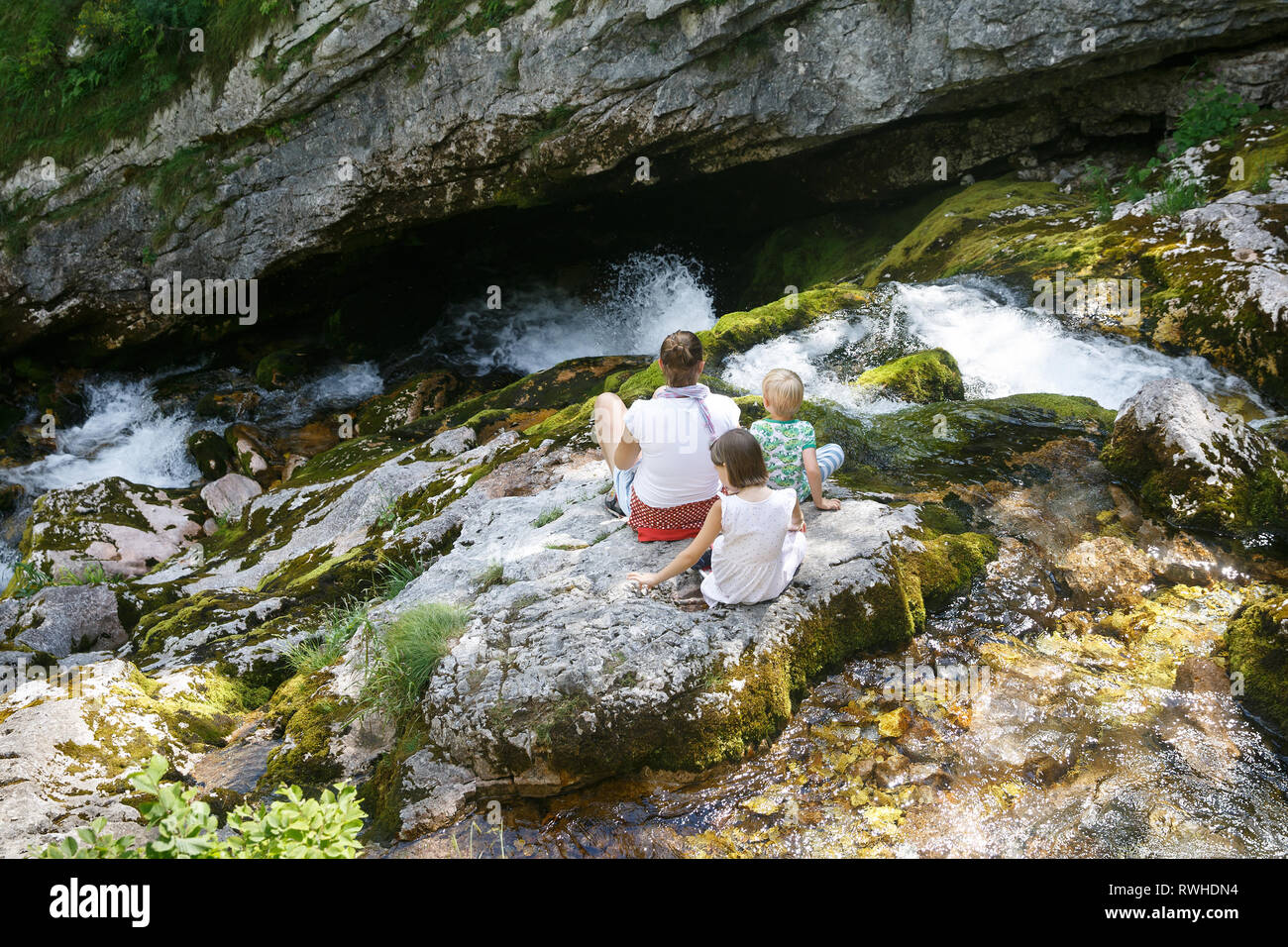 Mère avec des enfants assis sur les rochers par un ruisseau de montagne en été, de refroidissement. De vie de plein air, des parents, le concept de l'expérience de la petite enfance. Banque D'Images