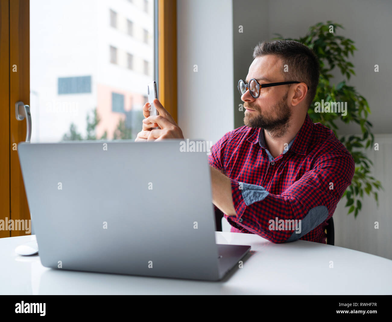 Accueil et communication concept - l'Homme à lunettes assis au bureau à domicile et de travailler sur un ordinateur portable et using smartphone Banque D'Images