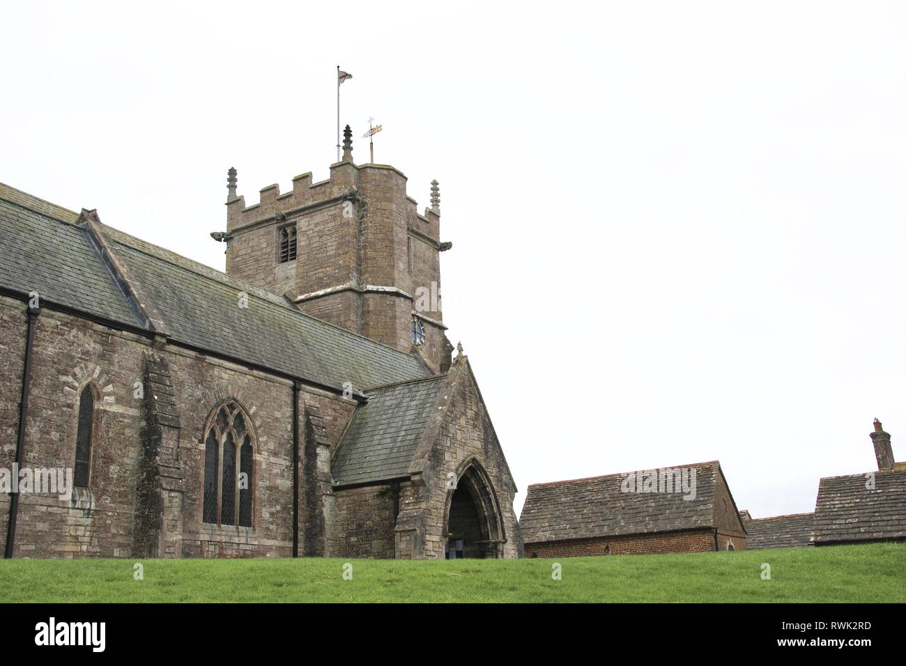 L'ancienne église de pierre de l'anglais (Norman) avec tour et flagstaff contre le ciel gris clair. Banque de l'herbe verte et toits d'immeubles, vue vers le haut Banque D'Images