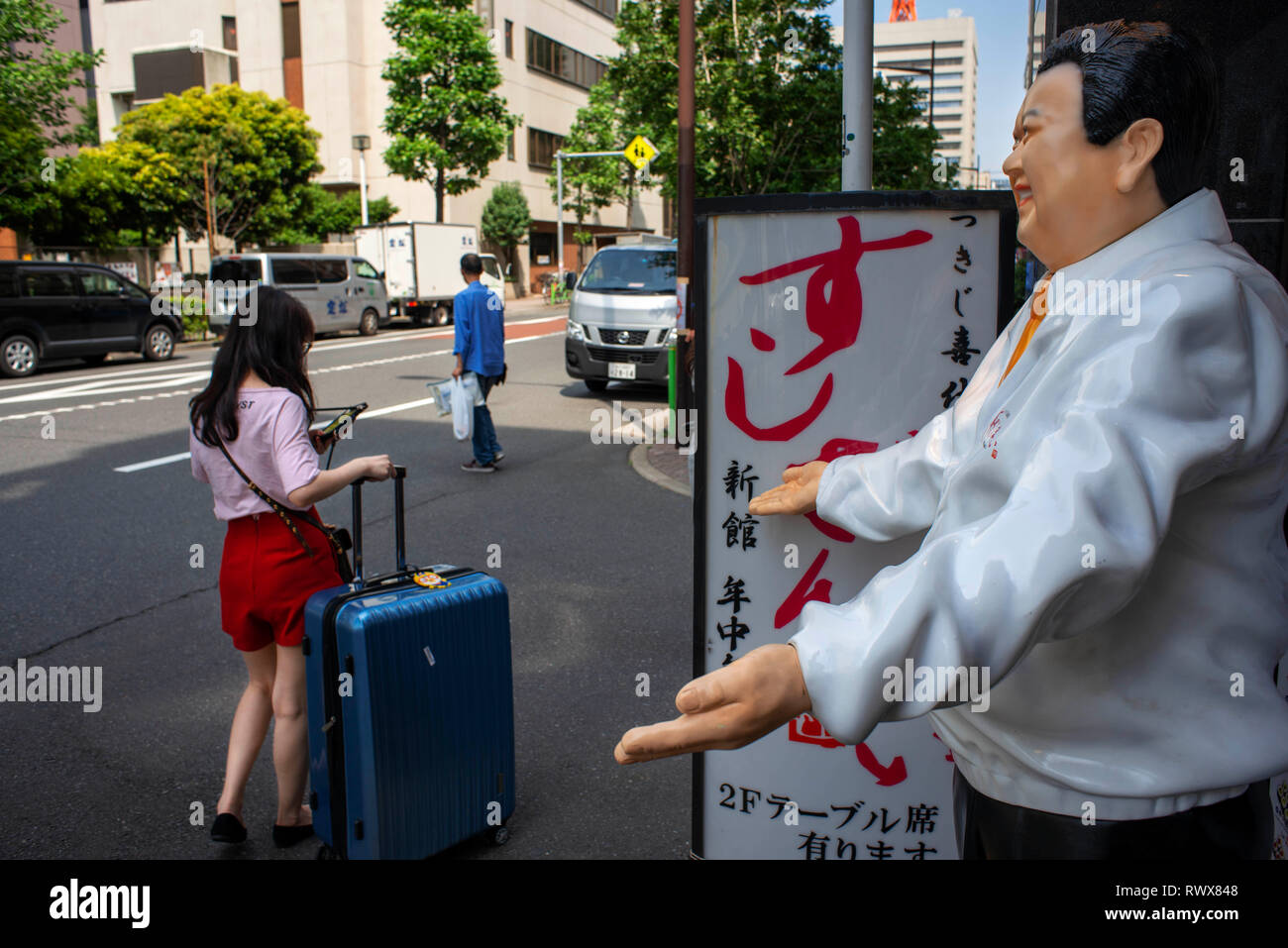 Scène de rue drôle à la porte du restaurant Sushi Terminal. Tokyo Japon. Un laissez-passer touristique locale avec une valise en face d'une statue de Kimura Kiyoshi, Banque D'Images