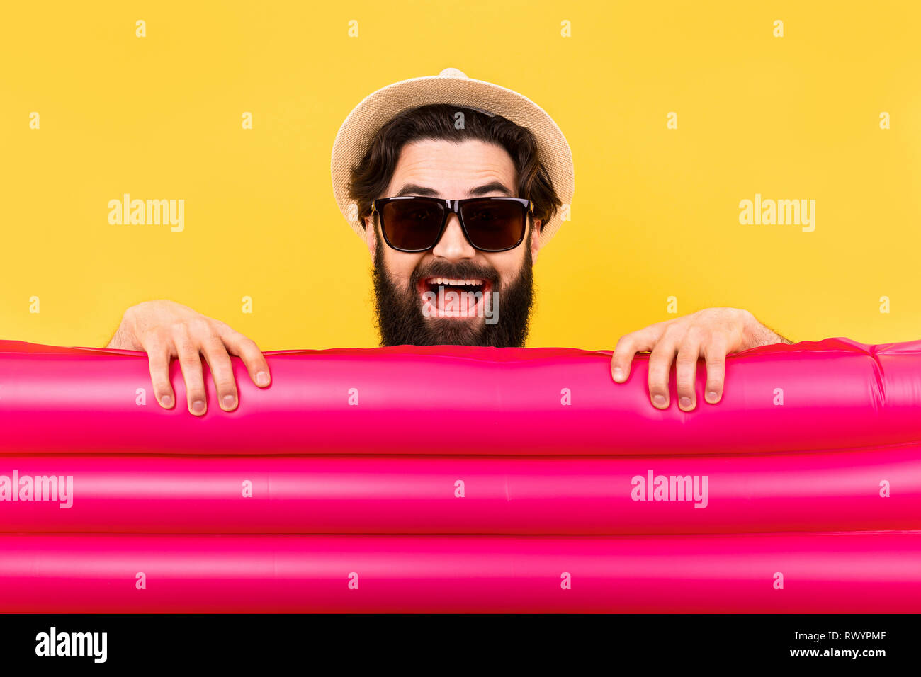 Studio portrait d'un barbu à lunettes de soleil et un chapeau de Panama, un homme tenant un matelas gonflable rose pour nager en face de lui, un concept summ Banque D'Images
