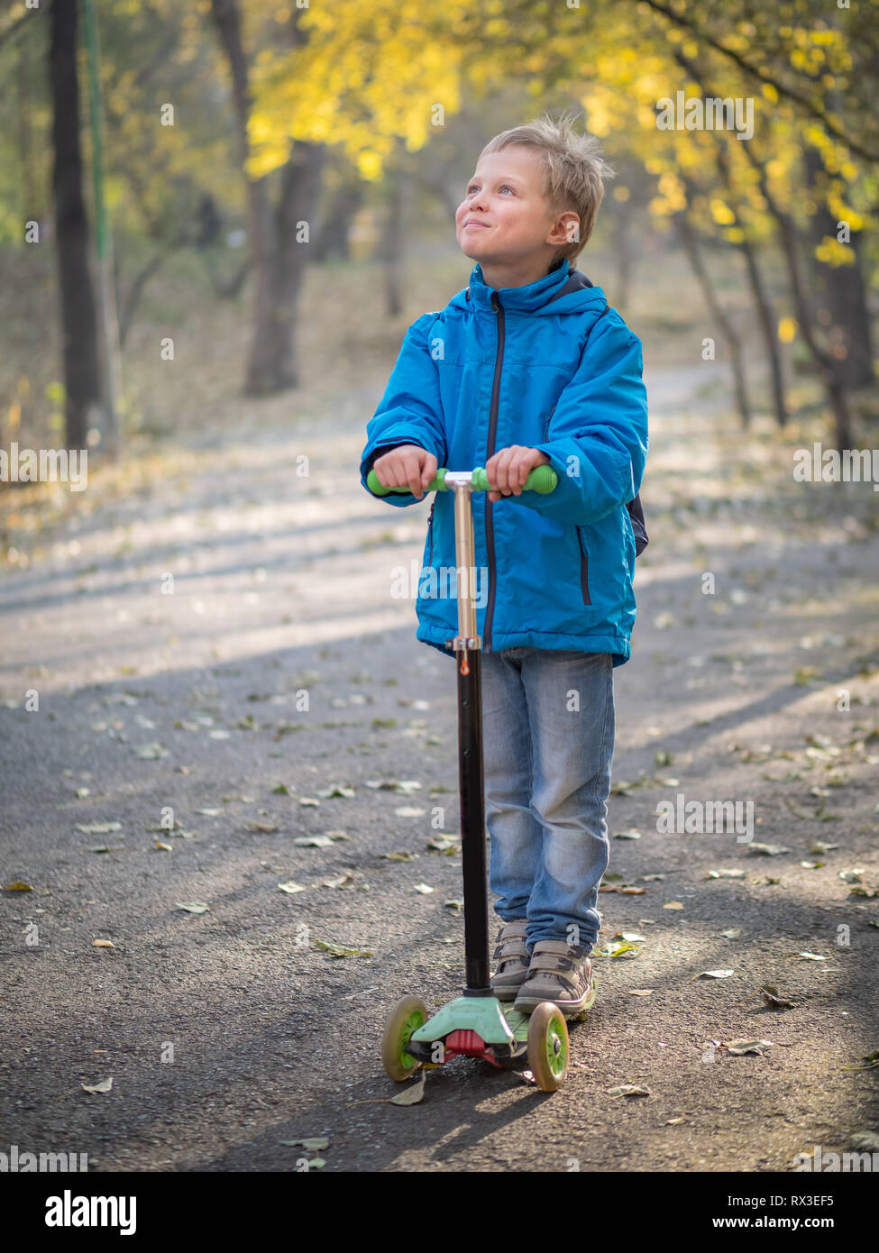 Un garçon en bleu vêtements équitation avec un sourire d'un scooter dans le parc de l'automne. Banque D'Images
