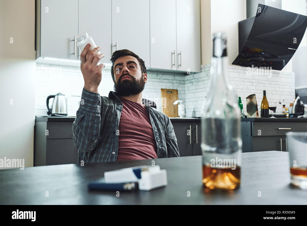 Dark-haired, triste et de gaspiller les boissons l'homme assis à la table, dans la cuisine, après avoir bu du whisky et à prendre de la drogue, complètement ivre, à la dep Banque D'Images