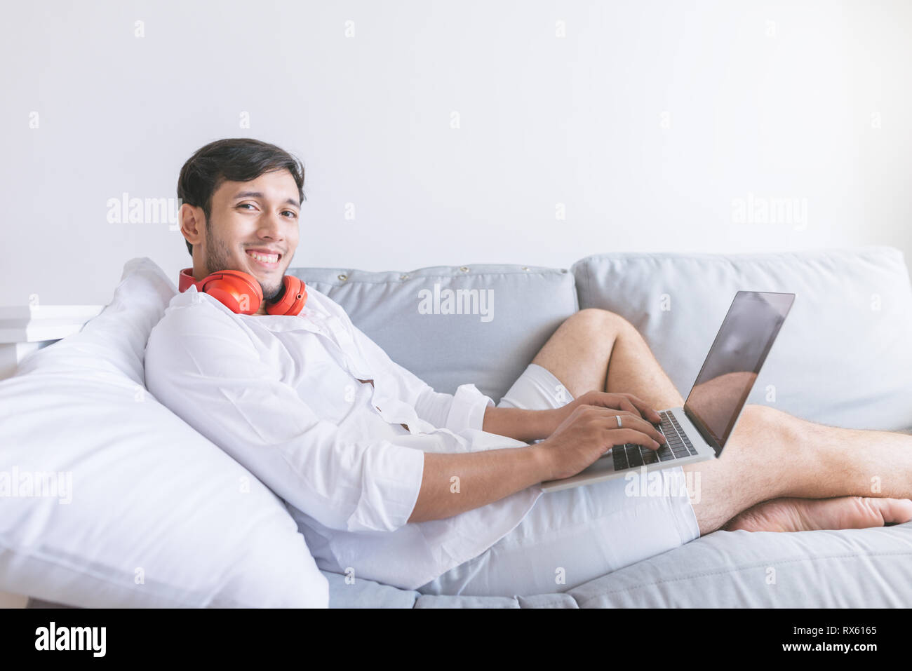 Jeune homme se détendre sur le canapé avec un ordinateur portable et un casque . Vue latérale du bel homme à l'aide de son ordinateur portable et à la recherche à l'appareil photo avec sourire tout en sitti Banque D'Images