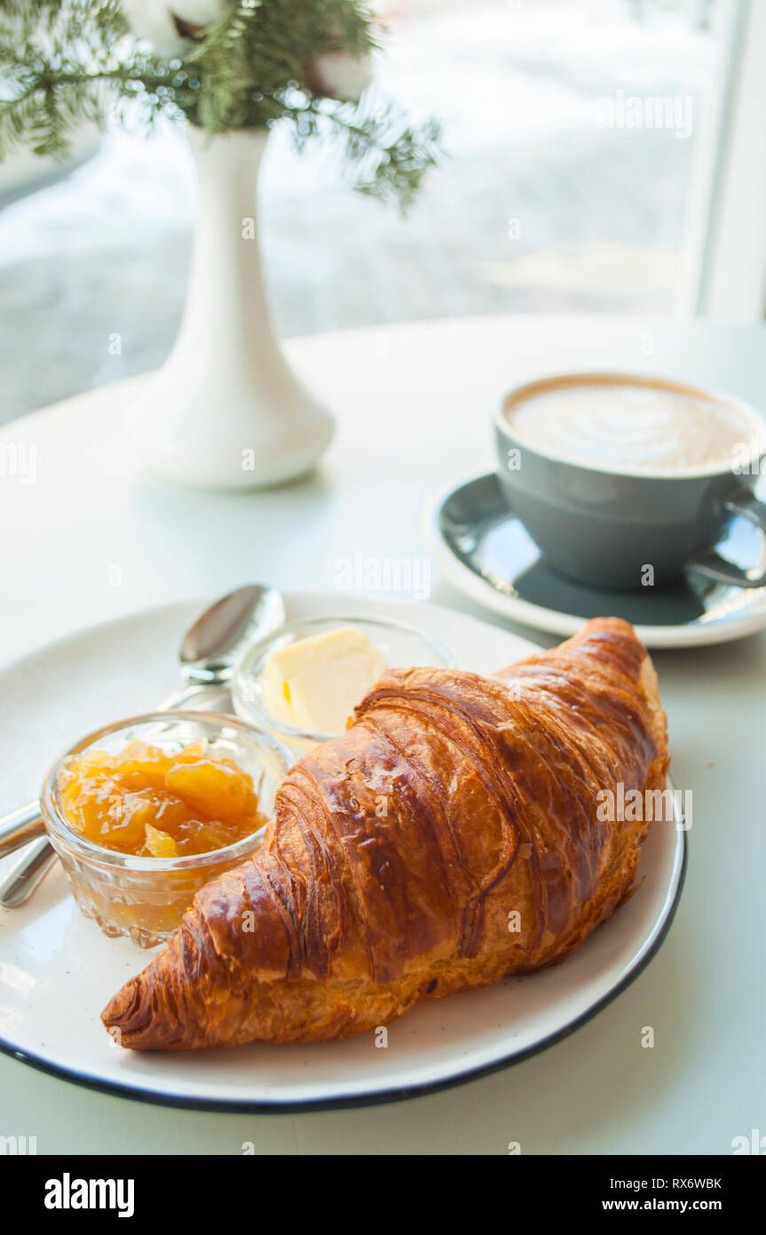 Croissant frais avec du beurre et de la confiture et du cappuccino avec belle latte art sur table gris dans un café. Banque D'Images