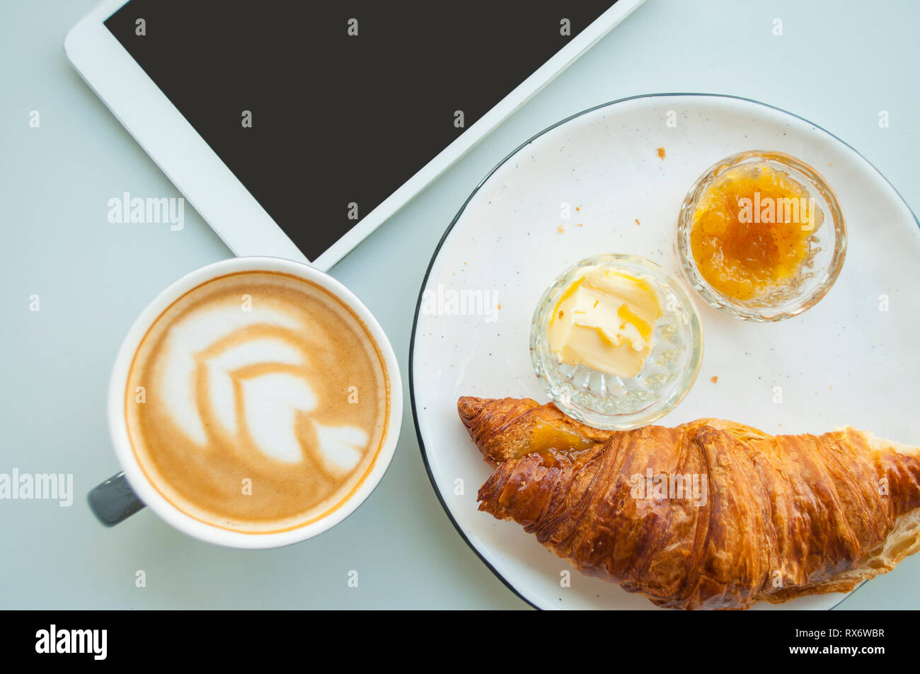 Croissant frais avec du beurre et de la confiture et du cappuccino avec belle latte art sur table gris dans un café. Mise à plat de style. Banque D'Images
