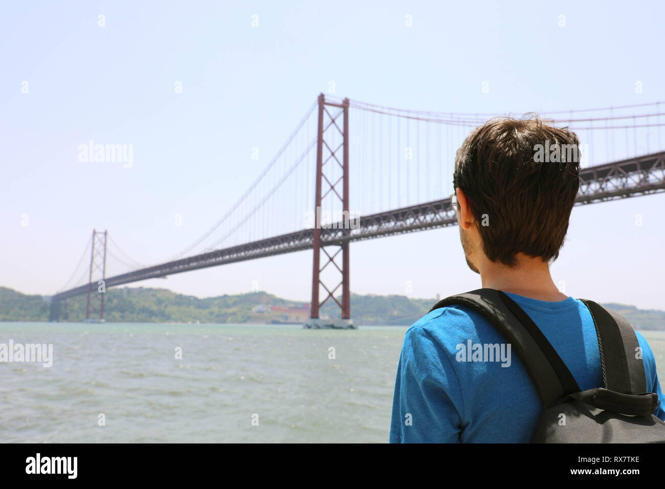 Vue arrière de jeune backpacker man looking at bridge. Voyageur ou touriste avec sac à dos sur le bord de l'eau à Lisbonne Portugal à côté du 25 avril Banque D'Images