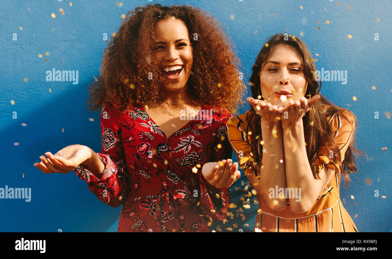 Les jeunes femmes attrayantes soufflant paillettes contre fond bleu. Mixed Race female friends having fun avec des paillettes. Banque D'Images
