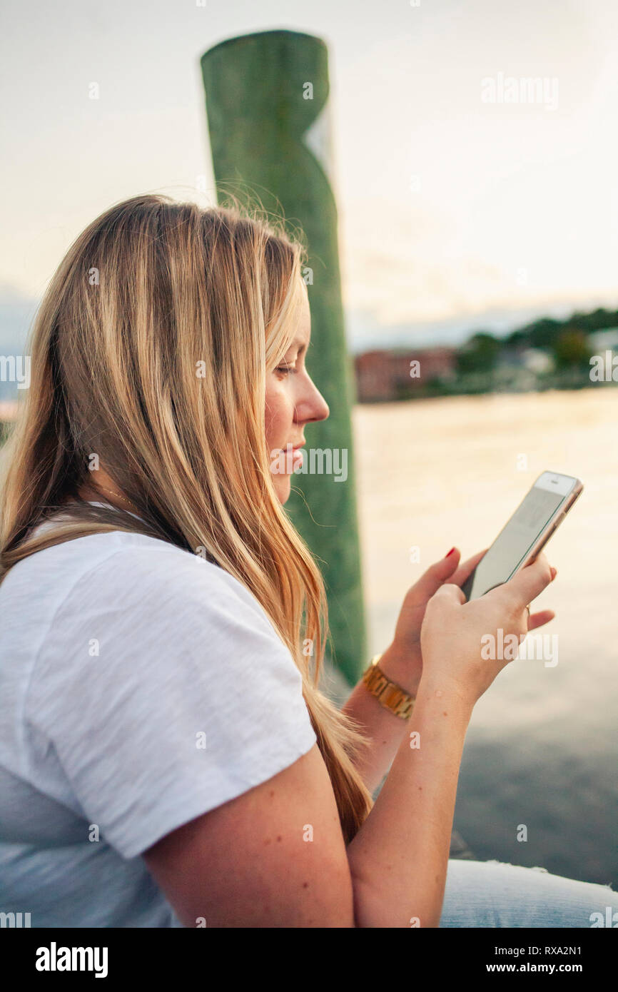 Vue latérale de la femme aux cheveux blonds à l'aide de smart phone while sitting at riverbank contre le ciel au coucher du soleil Banque D'Images