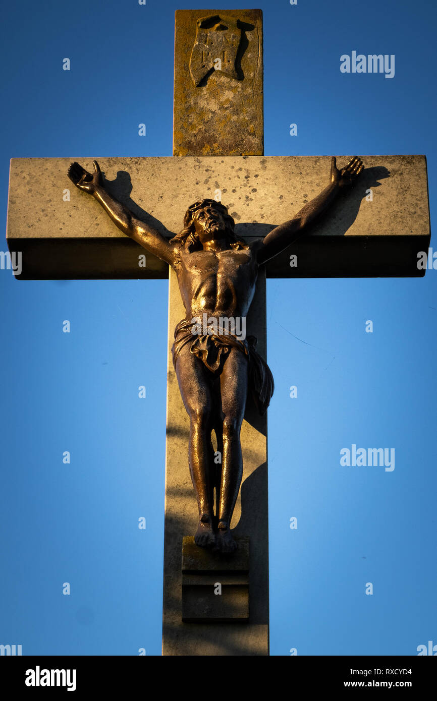 Belle croix en pierre crucifix avec Jésus Christ sculpture en bronze frappée par la lumière au coucher du soleil sur un ciel bleu. Banque D'Images