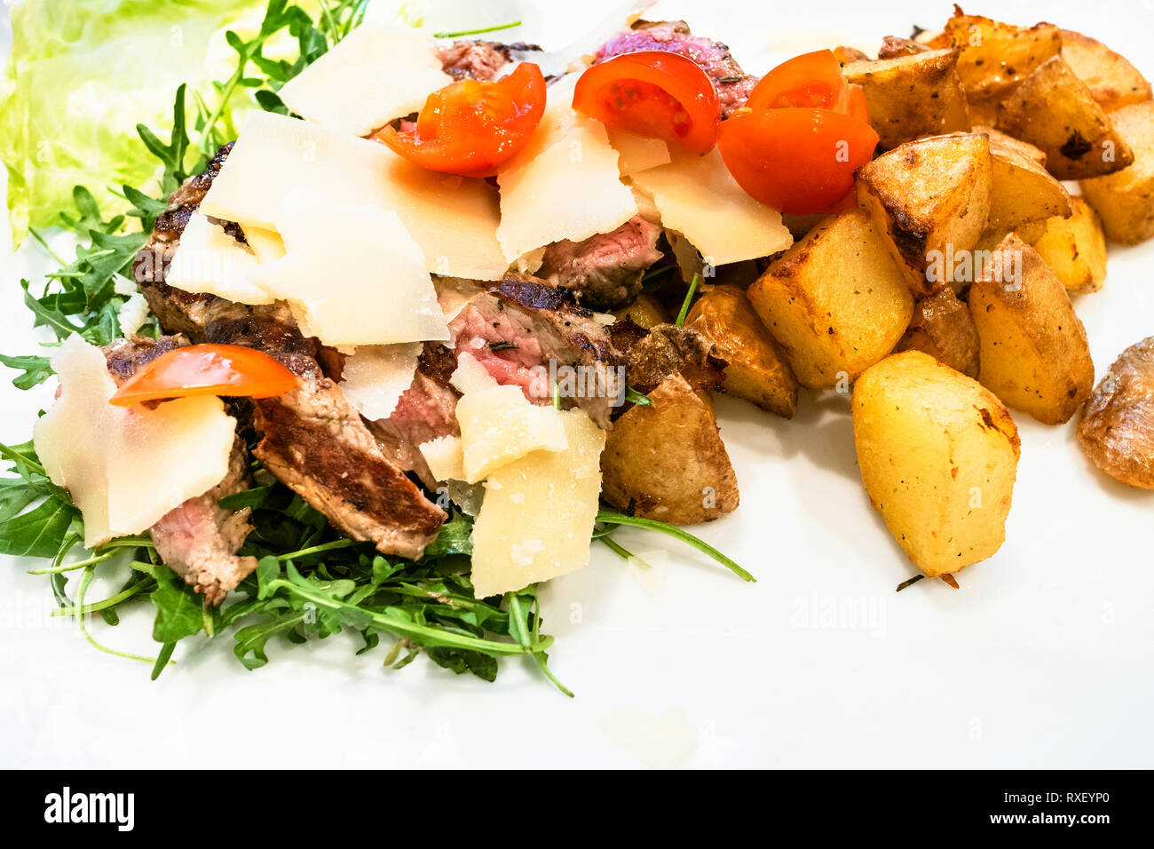 Cuisine italienne - Tagliata di manzo (tranches de rôti de bœuf angus rustique) avec pommes de terre rôties, fromage parmesan et salade de roquette close up on white plate Banque D'Images