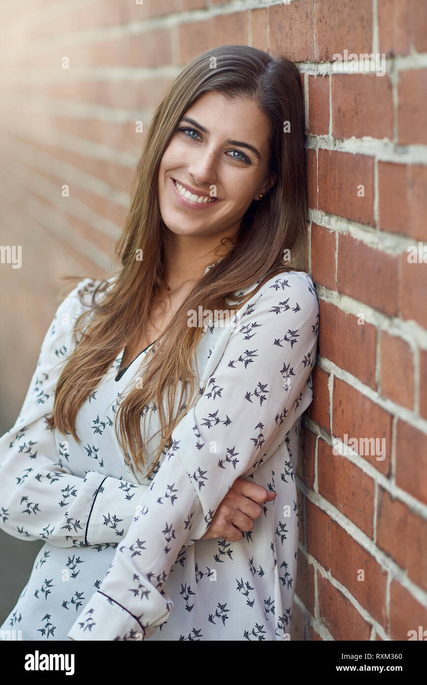 Jolie jeune femme avec un joli sourire naturel appuyé contre un mur de brique extérieur en se tournant vers la caméra avec flare sur le côté Banque D'Images