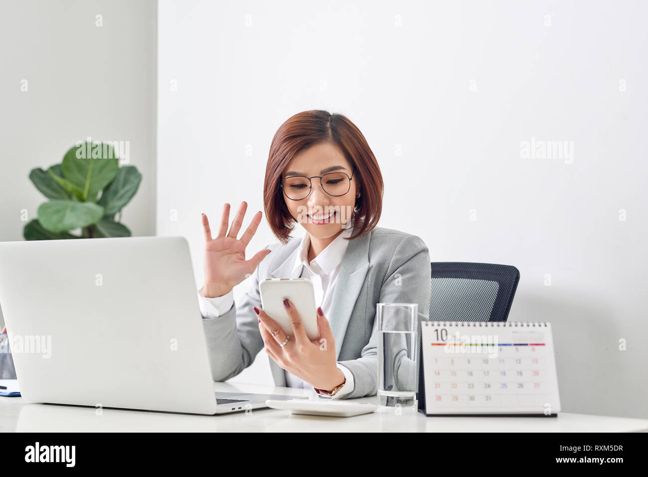Excitée femme font appel vidéo en agitant la main pour faire de vœux en tenant shot selfies sur téléphone mobile tout en s'asseoir au bureau de travail avec ordinateur portable isolé sur blanc b Banque D'Images