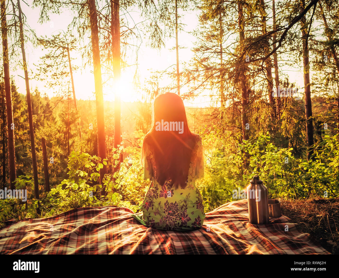 Belle jeune fille assise sur un plaid dans une clairière de la forêt au coucher du soleil autour de la beauté de la nature. À côté de la fille, c'est un thermos de fer Banque D'Images