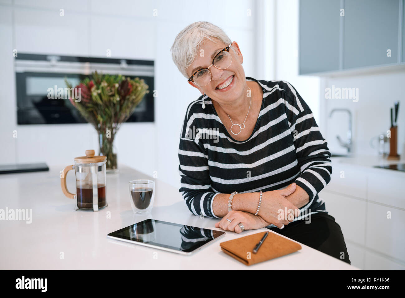 Portrait of a young woman smiling portant des lunettes et se penchant sur le comptoir de la cuisine. Senior woman working from home with tablet Banque D'Images