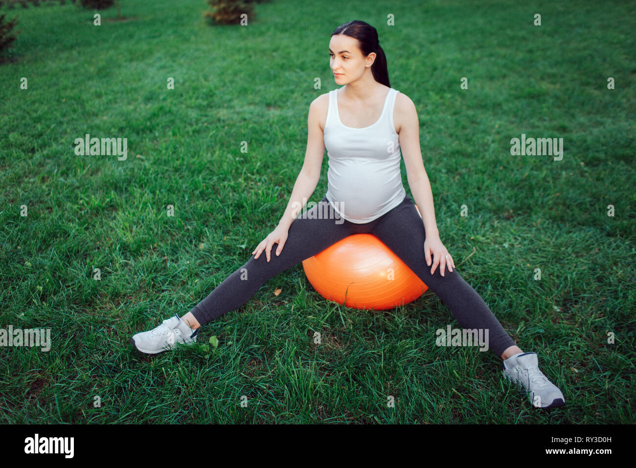 Jeune femme enceinte s'asseoir sur l'orange balle fintess avec jambes écartées en parc. Il voit à droite. Banque D'Images
