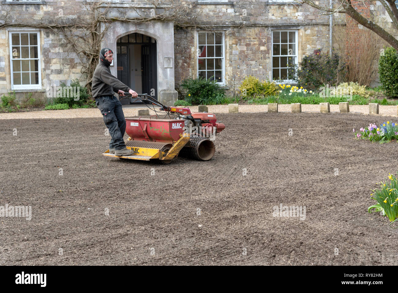 Winchester, Hampshire, Angleterre, Royaume-Uni. Mars 2019. Jardinier paysagiste à cheval sur un rouleau et de l'éparpilleur de semences pour former une nouvelle pelouse. Banque D'Images