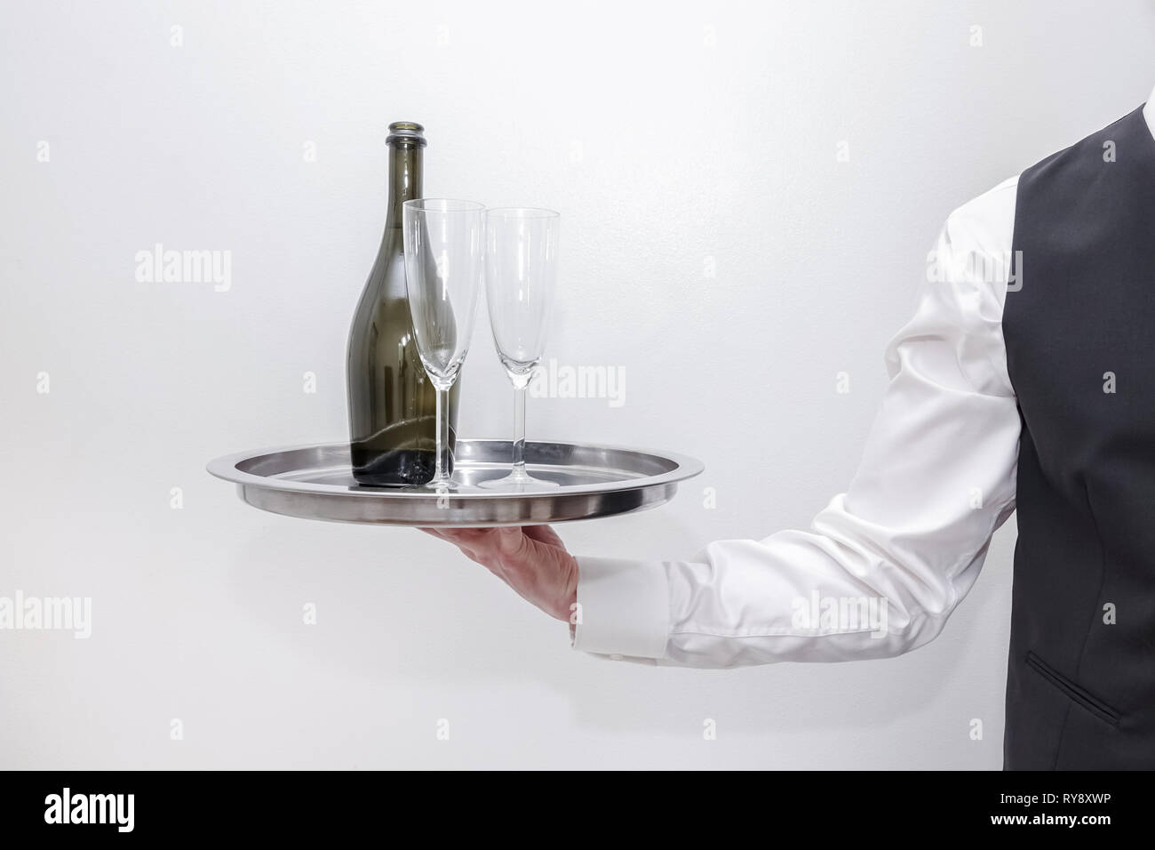 Un serveur / butler en chemise blanche et gilet costume noir portant un plateau d'argent avec une bouteille de champagne et deux verres. Arrière-plan blanc. Banque D'Images