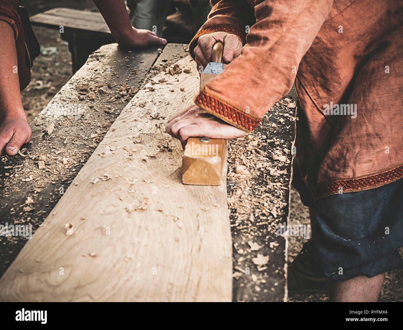 Carpenter à l'époque médiévale vêtements coton Travailler avec un bois en avion. L'homme s'accroche manuellement une planche en bois avec un avion. Construction d'une écorce de bois Banque D'Images