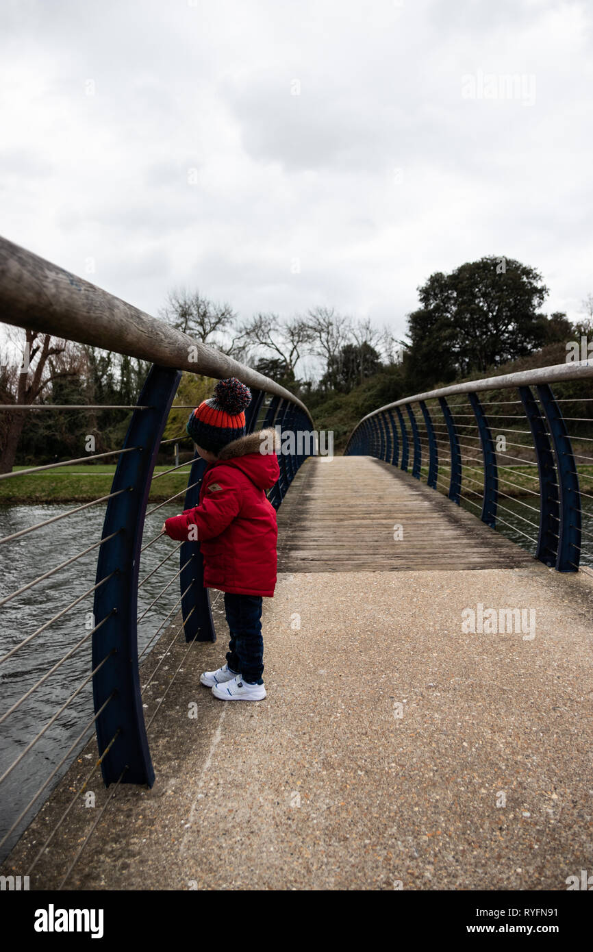 Deux ans en veste rouge et bobble hat sur un pont en bois à la recherche sur le côté au lac ci-dessous Banque D'Images