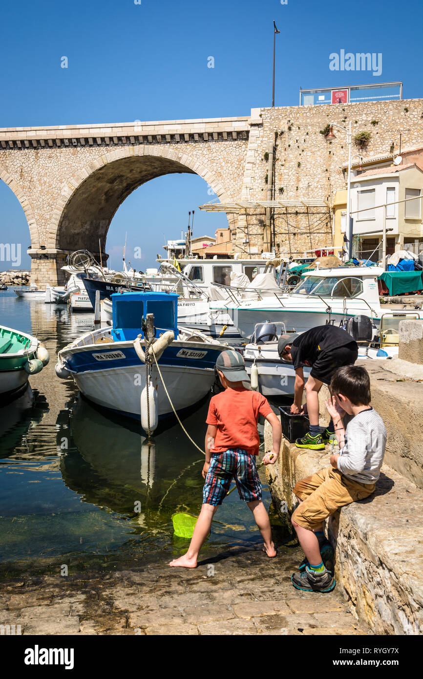 Trois garçons jouer près de la pêche bateaux dans le petit port du Vallon des Auffes à Marseille, France, avec le pont-route dans l'arrière-plan. Banque D'Images