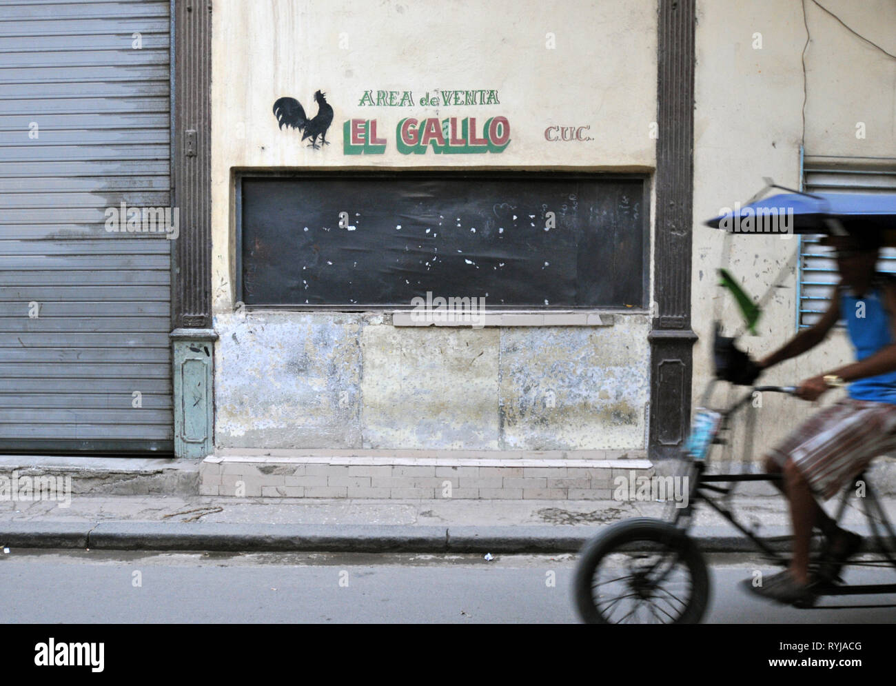 Un pedicab (vélo taxi) rides le long d'une route dans la Vieille Havane, Cuba, l'un des ventes au détail fenêtre avec un panneau espagnol El Gallo (Le Coq). Banque D'Images