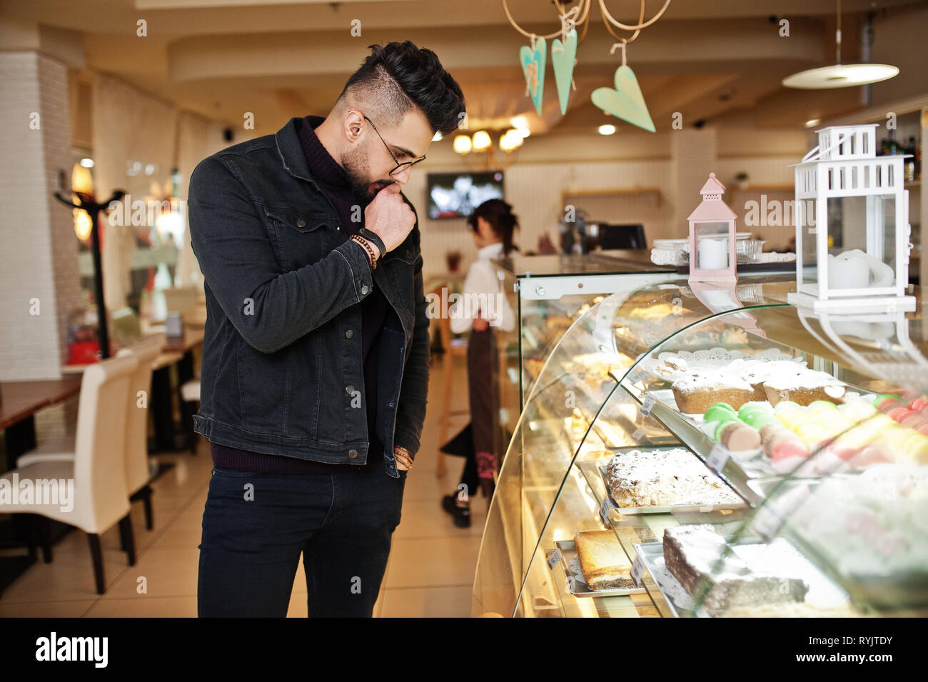 L'usure de l'homme arabes veste jeans noir et lunettes dans cafe choisir des gâteaux. À la mode et élégant modèle arabe guy. Banque D'Images