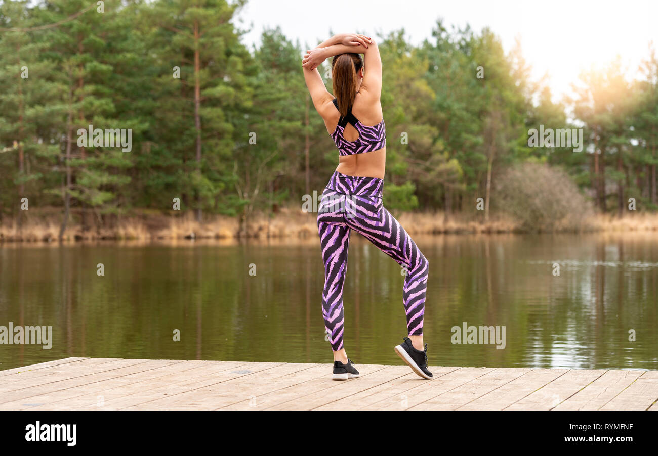 Sporty woman standing on une jetée par un lac chaud-up​ ce faisant les exercices. Vue arrière. Banque D'Images