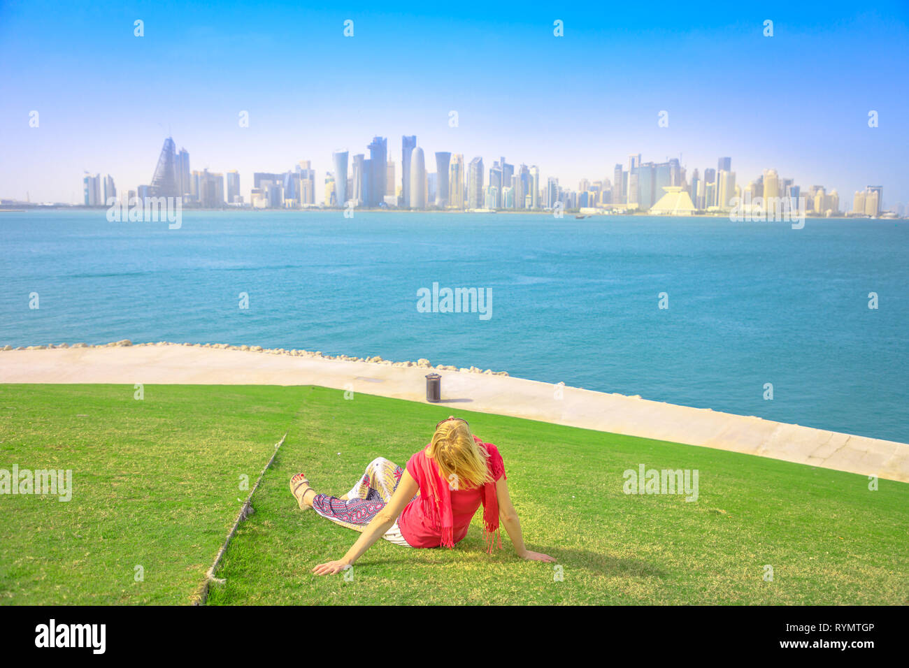 Caucasian woman sitting sur pelouse verte se penche sur les points de vue de West Bay à Doha au Qatar. Blonde touriste jouit d'Mound-Skyline d'ouest du centre-ville de Doha Banque D'Images
