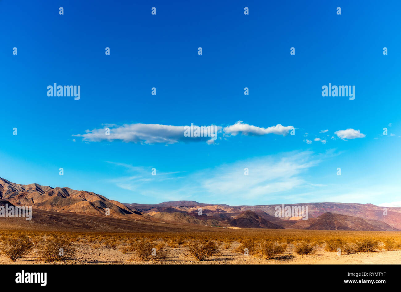 Lumière dorée sur la vallée du désert avec montagnes stériles au-delà sous un ciel bleu avec des nuages blancs. Banque D'Images