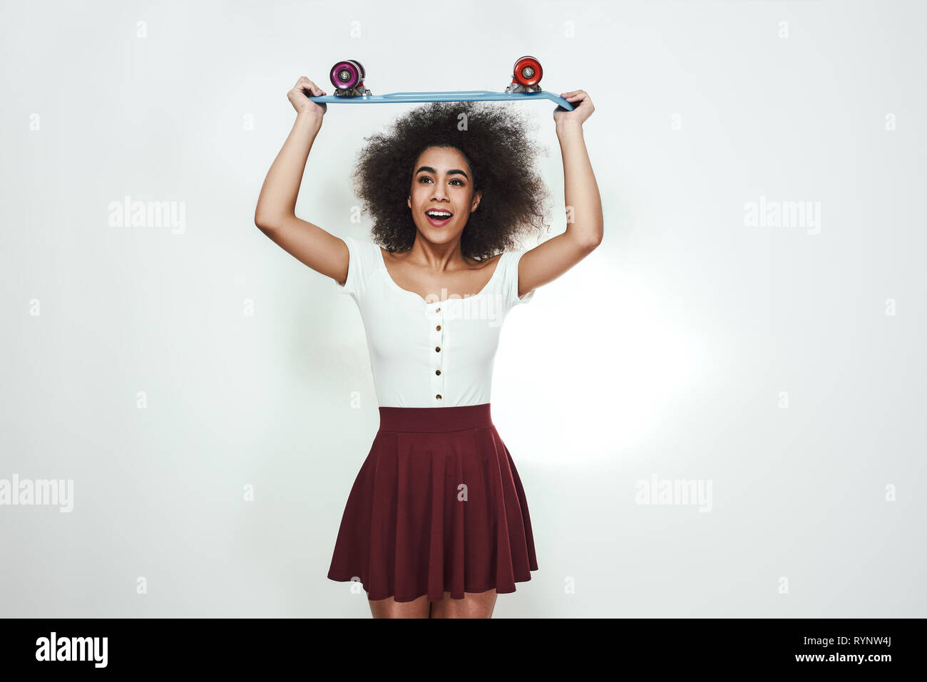 Profiter de la planche à roulettes. Portrait de jeune femme africaine ludique dans des vêtements décontractés holding skateboard sur sa tête et sourit tout en se tenant à l'arrière-plan gris. Mode de vie actif. Sport concept Banque D'Images