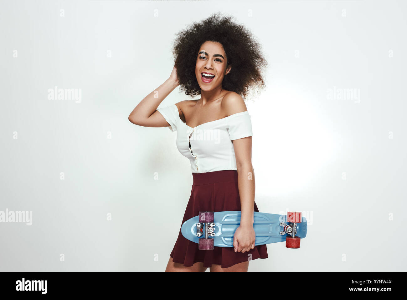 J'aime ma nouvelle planche à roulettes ! Jeune femme africaine avec des cheveux bouclés holding skateboard bleu et looking at camera avec sourire tout en se tenant contre un arrière-plan gris. Mode de vie actif. Sport concept Banque D'Images