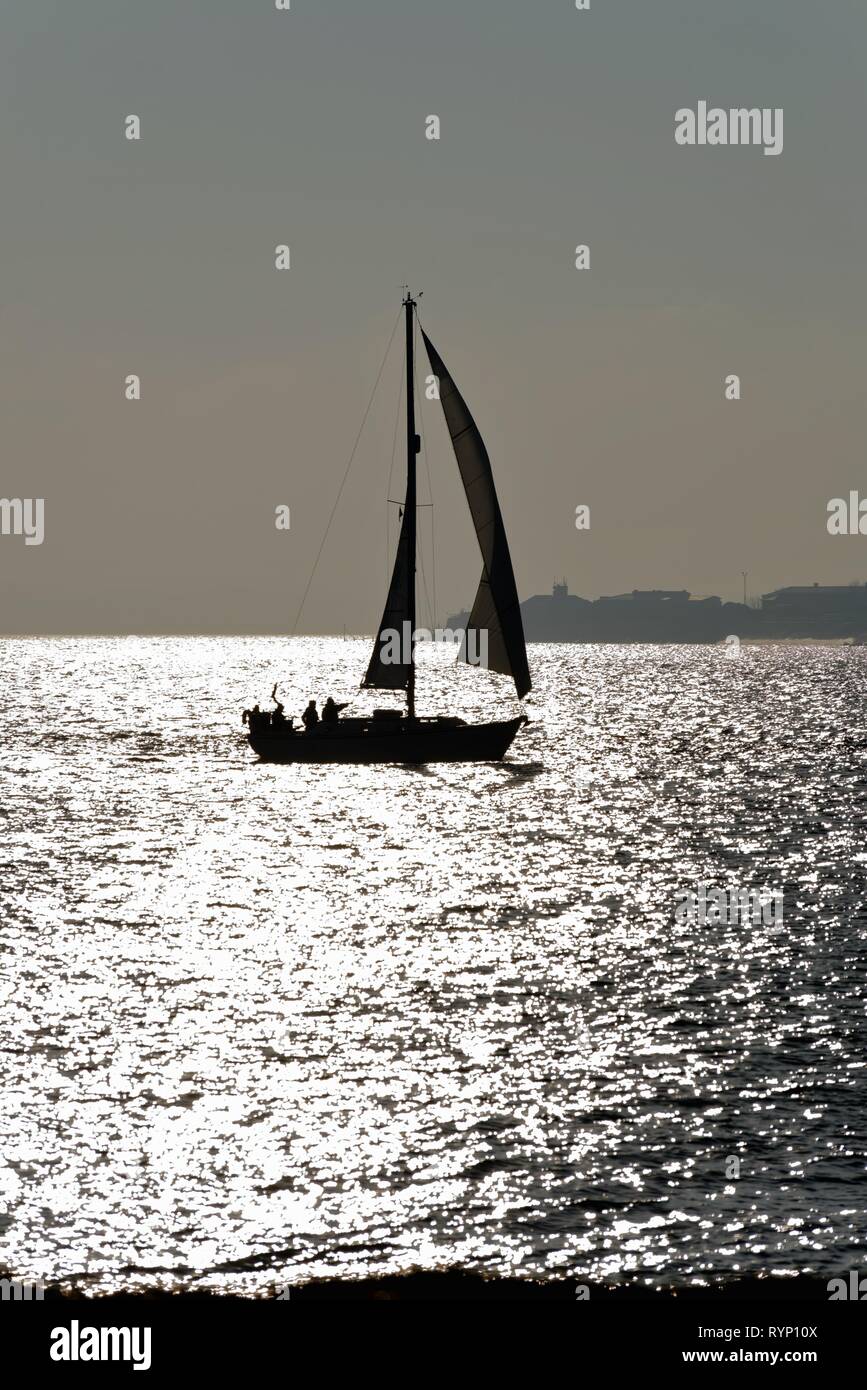 Silhouette d'un voilier naviguant sur le Solent à Portsmouth, Hampshire contre la réflexion de la lumière du soleil sur la mer, en Angleterre, Royaume-Uni Banque D'Images