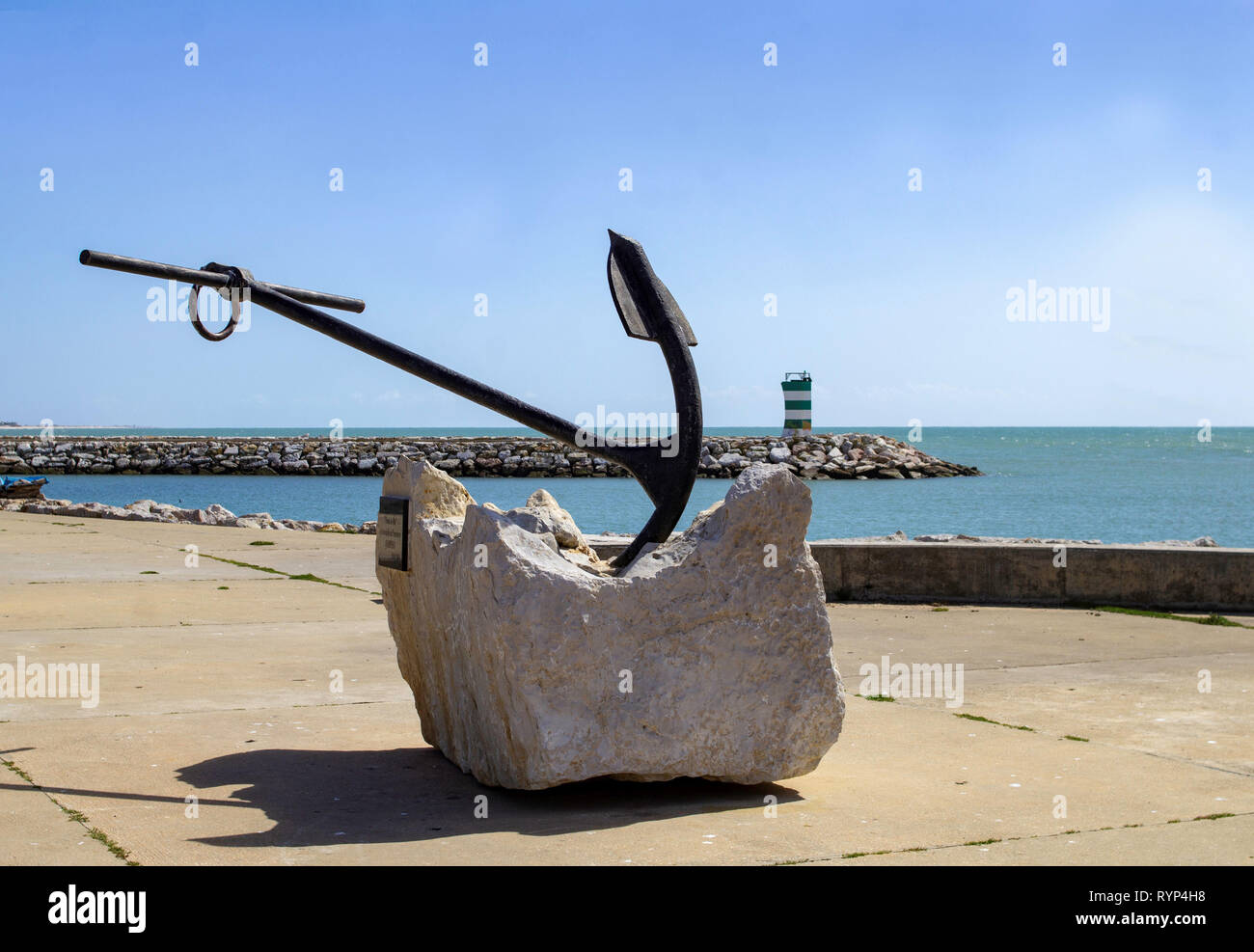 Quarteira, Portugal. Un ancien ancrage monté sur une base en béton avec un phare en arrière-plan. Banque D'Images