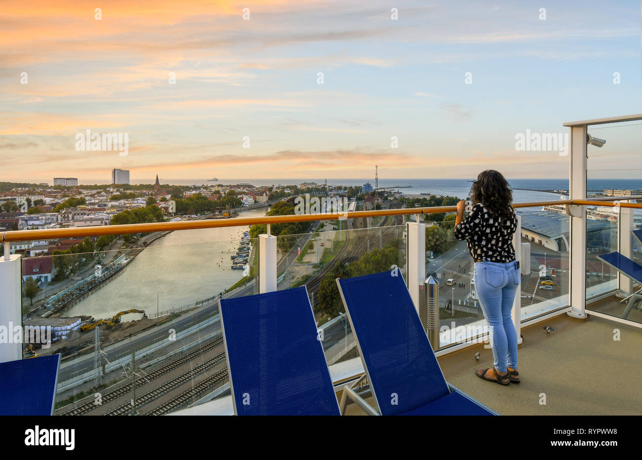 Une femme sur le pont supérieur d'un navire de croisière bénéficie d'coucher de soleil sur la ville côtière, plage, mer Baltique, Alter Strom canal et chemin de fer dans l'Allemagne de Warnemunde Banque D'Images