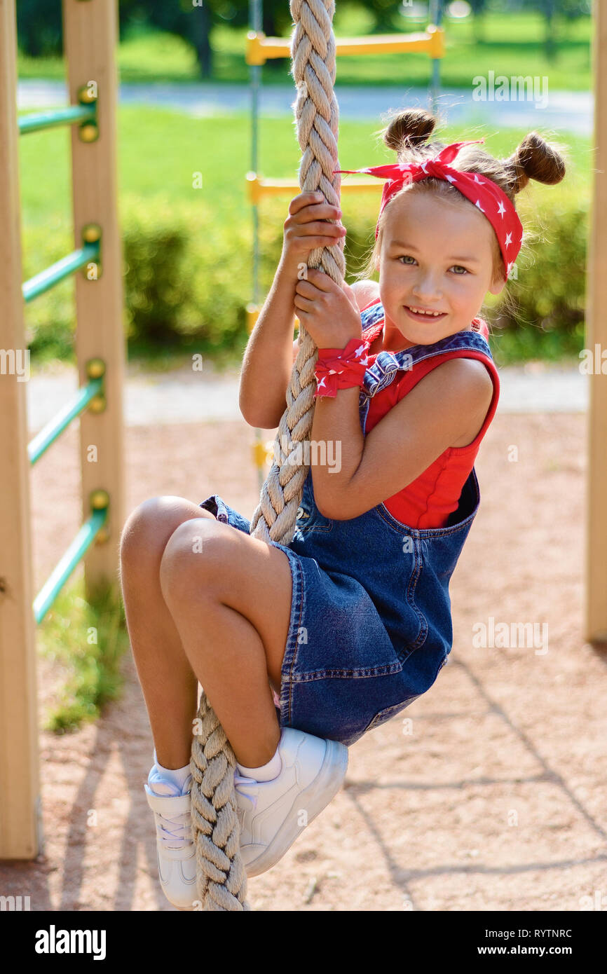Petite fille en salopette en jean et un T-shirt rouge et foulard joue et  s'amuser sur l'aire de jeux pour enfants. kid monter sur la corde Photo  Stock - Alamy