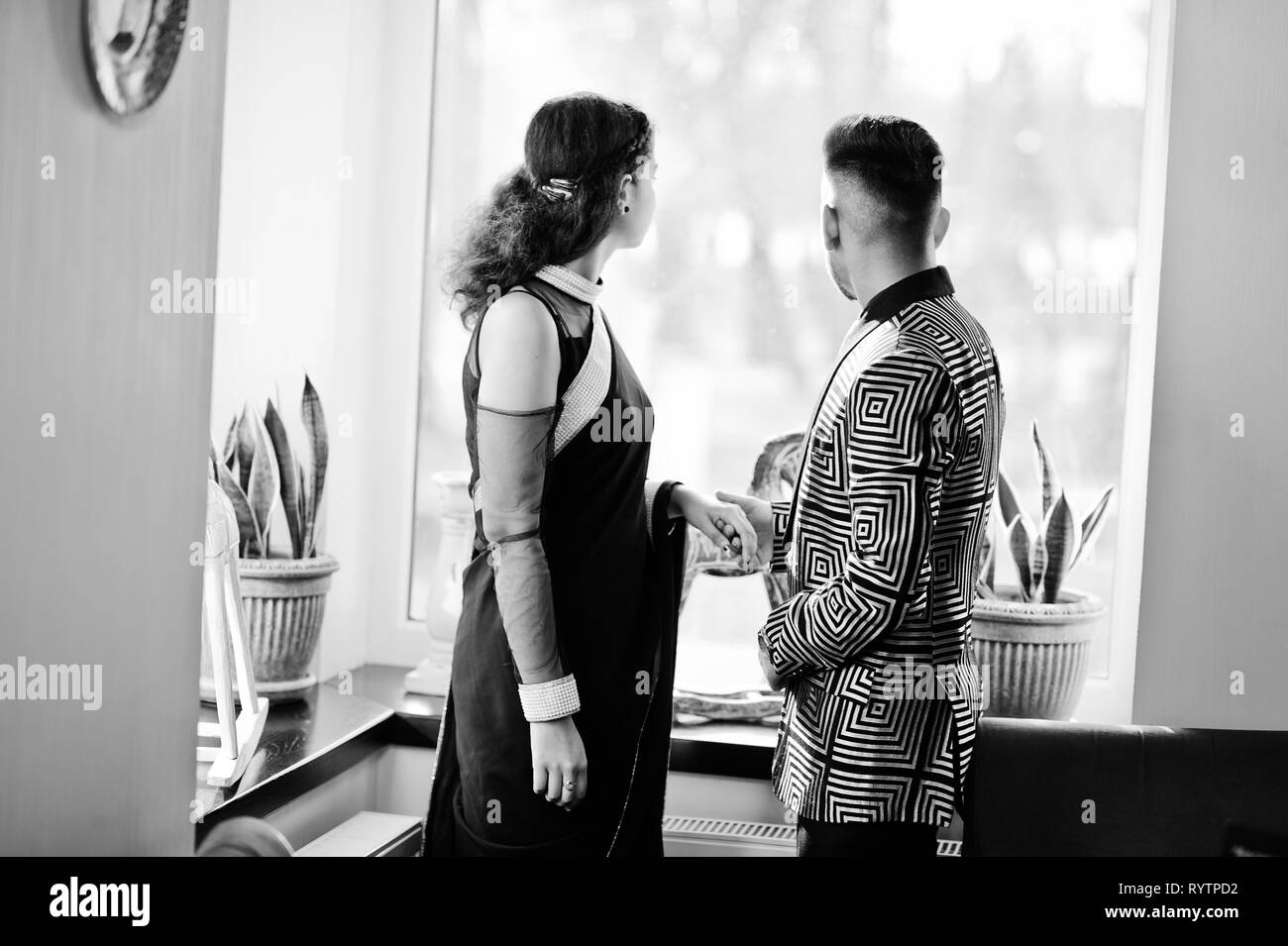 Joli couple indien dans l'amour, à l'usure et élégant costume, saree posés sur un restaurant. Retour d'entre eux, ils ont à la recherche sur la fenêtre et détient les mains. Banque D'Images
