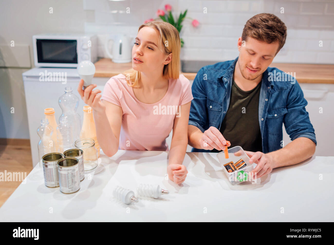 Le recyclage, la réutilisation. Jeune couple batteries tri, ampoules dans de petits récipients avec symbole recyclage assis à cuisine Banque D'Images