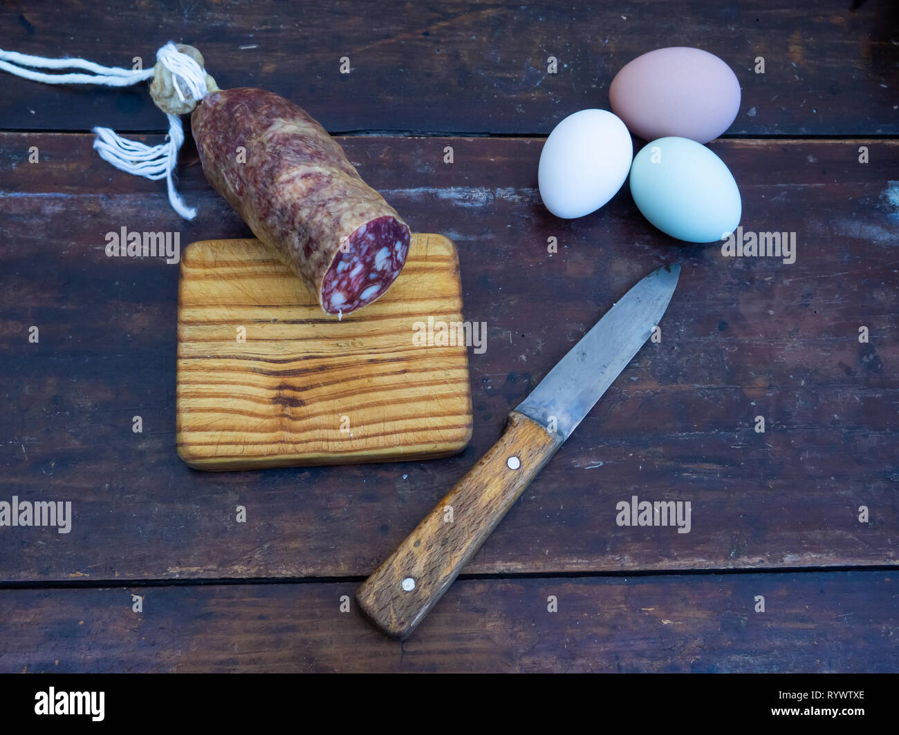 Une saucisse de porc ibérique sur une vieille planche en bois avec un ancien couteau et trois oeufs multicolores Banque D'Images