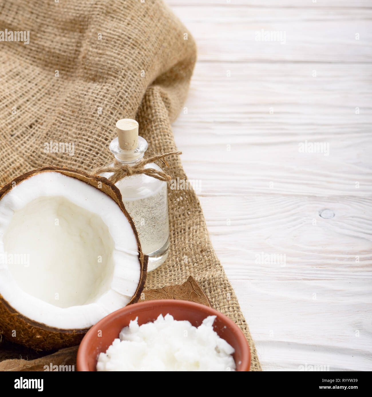 L'huile de noix de coco dans un bol d'argile brune, bouteille de verre, coque de noix de coco avec de la viande sur le chanvre sacs sur une table de cuisine en bois blanc Banque D'Images