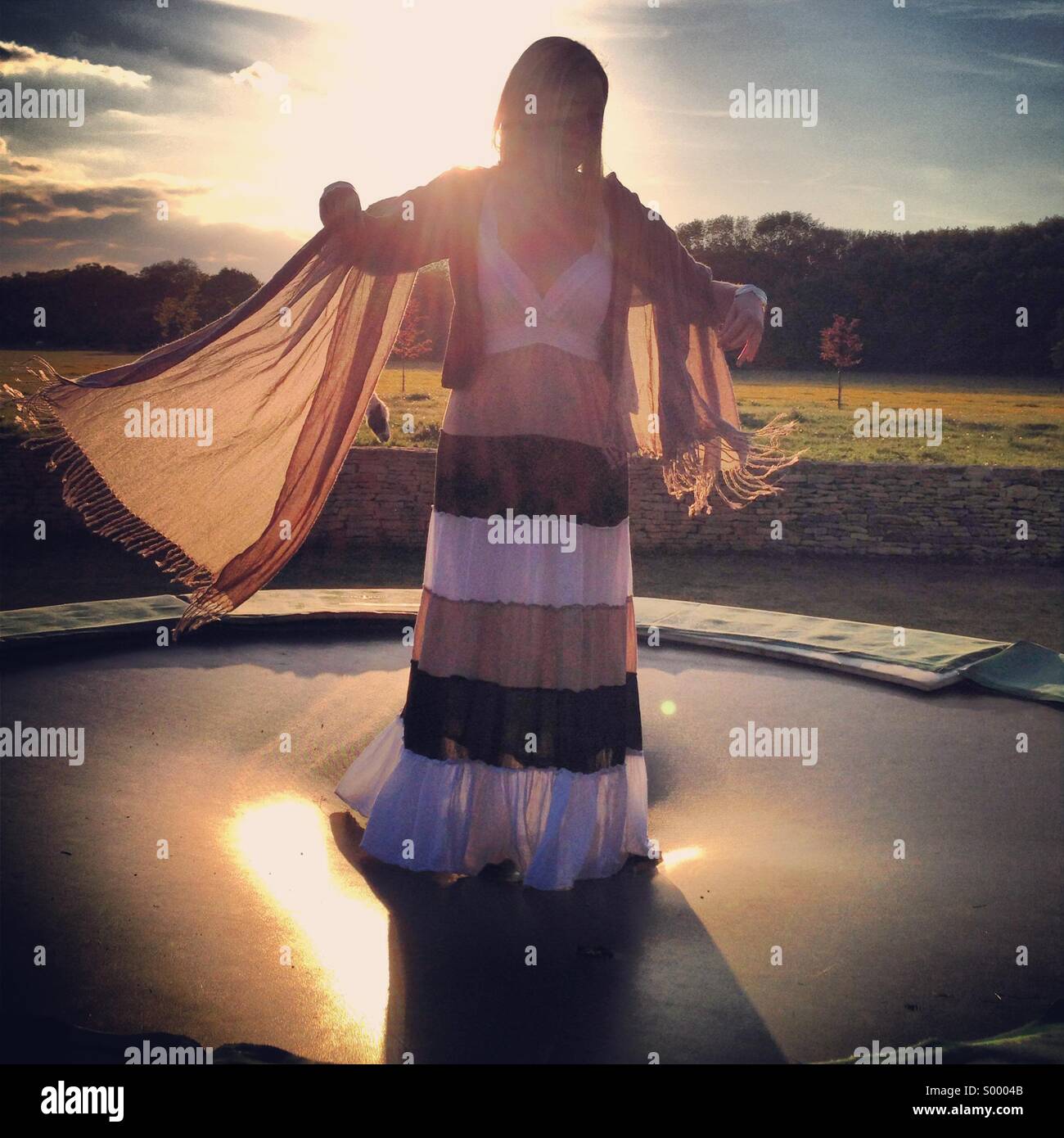 Une jeune femme en robe flottant rebondit sur un trampoline au coucher du soleil. Banque D'Images