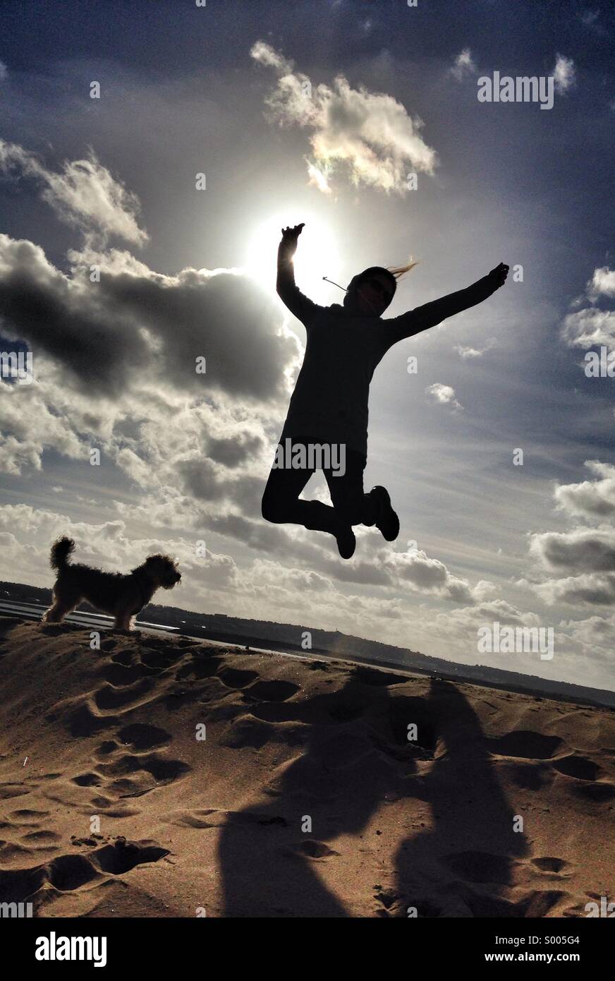 Une femme saute de joie sur une journée ensoleillée à la plage, dans le Devon, Royaume-Uni. Beau ciel bleu et nuages blancs moelleux sont vus dans l'arrière-plan comme la dame est découpé sur le soleil Banque D'Images