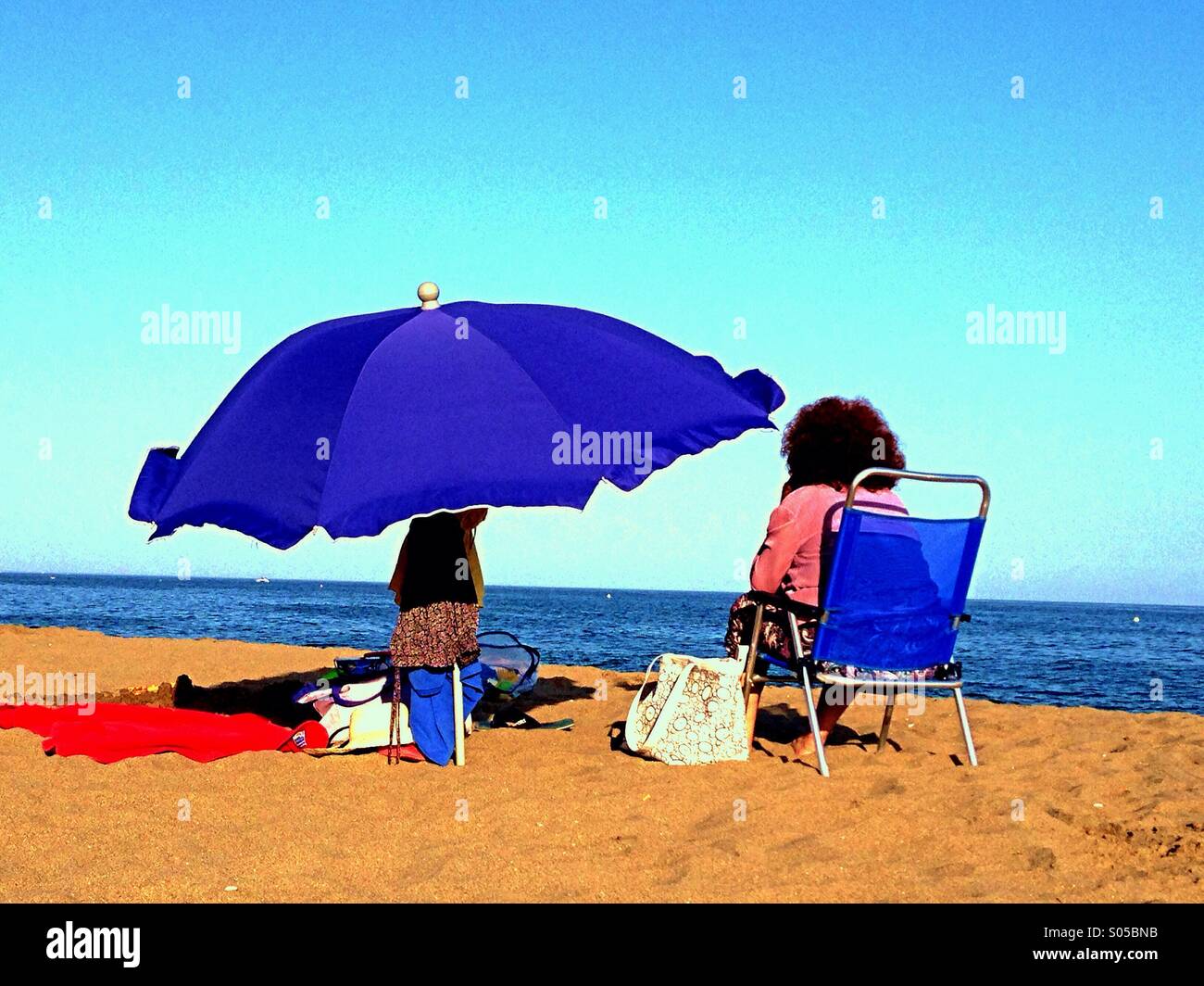 Une vieille femme avec les cheveux crépus est assis tout habillé sur une chaise  longue sur une plage dans le soleil de plomb à côté d'un parasol. La plage  est à La
